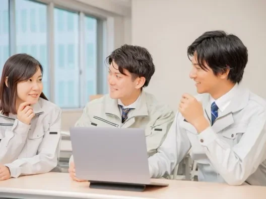 ノートパソコンを囲んで談笑する作業着姿の3人のスタッフ